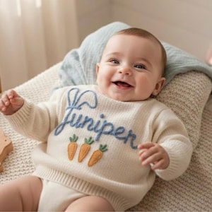 May include: A baby wearing a cream-coloured jumper with the name "Juniper" embroidered in blue script. The jumper features three orange carrot appliqués with green tops. The baby is lying on a cream-coloured blanket.