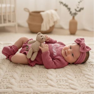 May include: A baby in a mauve knitted outfit, comprising a long-sleeved onesie, booties, and a headband with a bow. The outfit is embroidered with the name "Quinn". The baby holds a small, beige teddy bear.