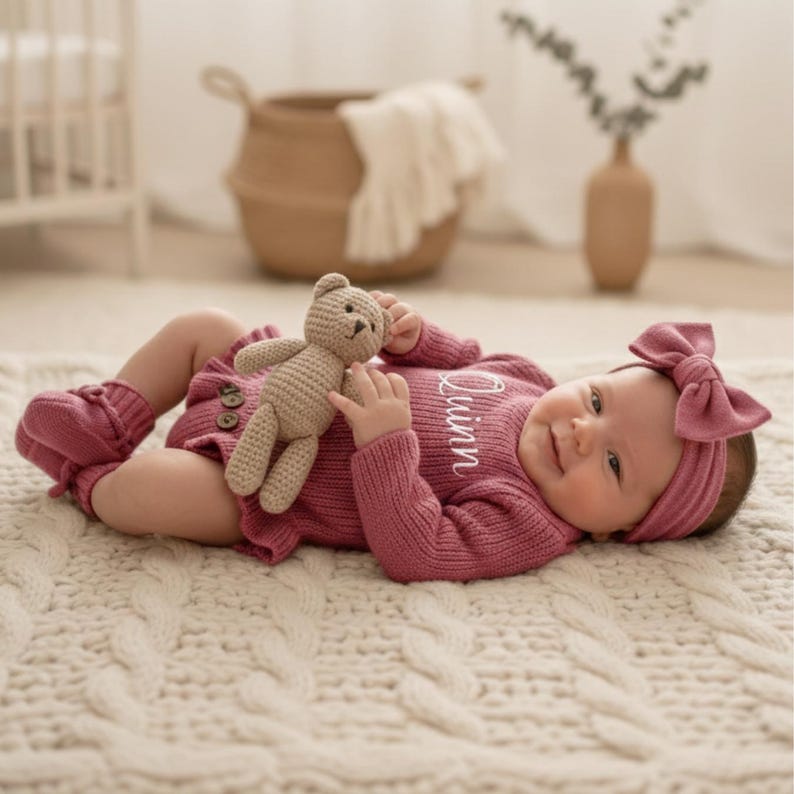 May include: A baby in a mauve knitted outfit, comprising a long-sleeved onesie, booties, and a headband with a bow. The outfit is embroidered with the name "Quinn". The baby holds a small, beige teddy bear.
