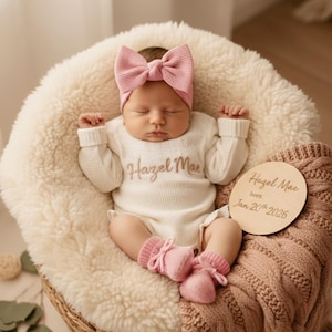May include: A baby wearing a white knitted onesie with the name "Hazel Mae" embroidered on it, a pink bow headband, and pink booties. The baby is lying on a fluffy white cushion. A wooden disc with the baby's name and birthdate is visible.