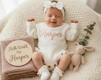 May include: A baby wearing a white onesie embroidered with the name "Harper" in pink, a matching headband, and socks. A wooden sign with the text "Hello World. I'm Harper" is next to a pink blanket. A soft toy rabbit is also present.