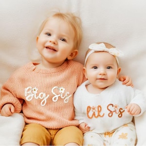 May include: Two babies wearing matching knit sweaters with the words "Big Sis" and "Lil Sis" embroidered on them. The sweaters are peach and white with floral accents. The babies are smiling and looking at the camera.