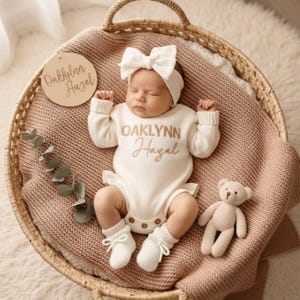 May include: A baby wearing a white outfit with the name "Oaklynn Hazel" embroidered on it, a matching bow headband, and socks, lies in a woven basket. A small teddy bear and a wooden sign with the same name are also in the basket.