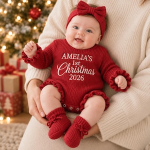 May include: A red knitted long-sleeve romper with ruffled cuffs and leg openings, featuring the white embroidered text AMELIA'S 1st Christmas 2026. The outfit includes a matching red knitted headband with a bow and red knitted baby booties.