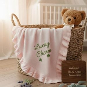Könnte beinhalten: Ein Weidenkorb enthält eine rosa Babydecke mit gerüschtem Rand und den grün gestickten Worten "Lucky Charm" sowie zwei Kleeblättern. Ein brauner Teddybär mit weißer Schleife sitzt im Korb. Ein Holzschild lautet "Willkommen Baby Anna - Januar 2026."