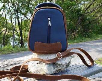 May include: A small blue leather backpack with a brown leather trim and a long brown leather strap. The backpack has a zipper closure and is sitting on a rock.