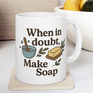 Pode incluir: Caneca de cerâmica branca com o texto "When in doubt, Make Soap" em letras marrons. A caneca apresenta ilustrações de ingredientes para fazer sabão. A caneca está sobre um porta-copos bege.