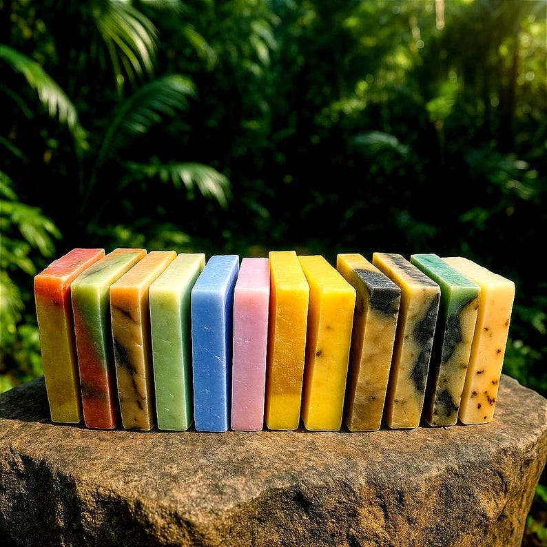 May include: A row of 12 colorful bars of soap arranged on a rock surface. The soap bars are in shades of green, blue, yellow, orange, and pink.