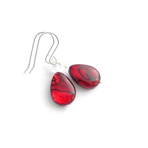 May include: A pair of teardrop-shaped earrings with a deep red, marbled appearance. The earrings have silver-colored ear wires and small clear crystal accents. The background is plain white.