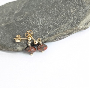 May include: Gold stud earrings featuring red and brown speckled gemstone beads. The earrings are presented on a grey stone surface against a white backdrop. The earrings have a post and butterfly backing.