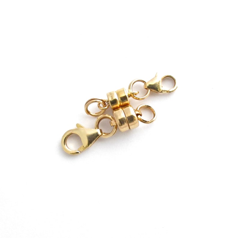 May include: Gold magnetic clasp for a necklace or bracelet. The clasp has two round magnetic pieces connected by a chain with two lobster claw clasps.