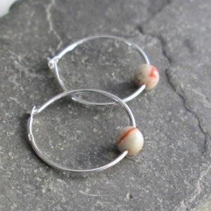 May include: A pair of silver hoop earrings. Each earring features a small, round, beige bead with a red stripe. The earrings are displayed on a gray, textured surface.