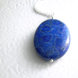 May include: Oval, blue pendant with a floral pattern, suspended from a silver chain. The pendant has a polished, glossy finish. The background is a textured white surface.