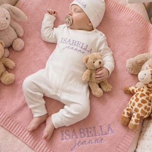 Może przedstawiać: Noworodek w białym pajacyku i czapce, oba z haftem "Isabella Jeanne", leży na różowym, dzianinowym kocyku. Pluszowe zabawki, w tym miś, królik i żyrafa, otaczają dziecko. Karuzela wisi nad łóżeczkiem.