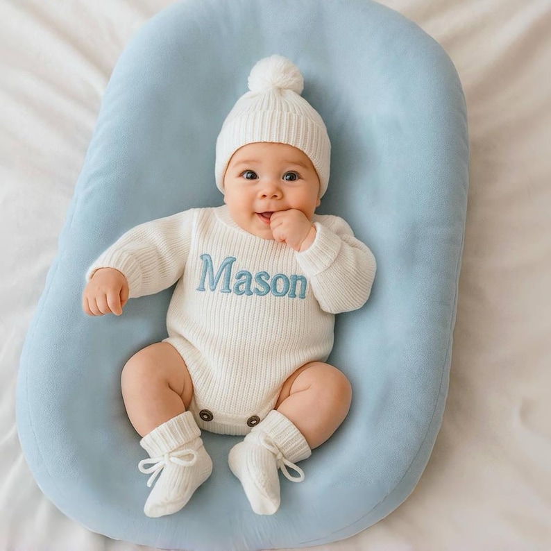 Może przedstawiać: Niemowlę w białym dzianinowym stroju, w tym czapka z pomponem, body z długim rękawem z wyhaftowanym na niebiesko imieniem "Mason" i pasujące skarpetki z kokardkami. Dziecko leży na jasnoniebieskiej, wyściełanej powierzchni.