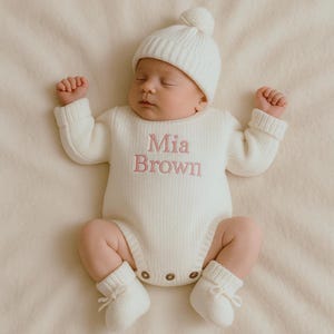 Może przedstawiać: Noworodek w białym, dzianinowym stroju, w tym czapka z pomponem, body z długim rękawem i buciki. Imię "Mia Brown" jest wyszyte na r&oacute;żowo na body. Dziecko leży na kremowym kocu.