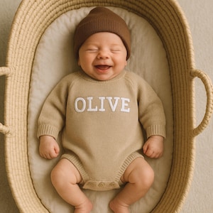 Może przedstawiać: Uśmiechnięte dziecko w beżowym dzianinowym śpioszkach z napisem "OLIVE" białymi literami. Dziecko ma również na sobie brązową czapkę. Dziecko leży w wiklinowym koszu.