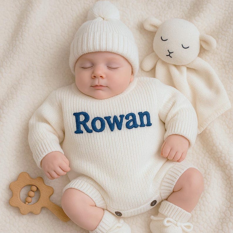 Może przedstawiać: Niemowlę w kremowym, dzianinowym śpioszku z wyhaftowanym na granatowo imieniem "Rowan". Dziecko ma r&oacute;wnież pasującą dzianinową czapkę i buciki. W pobliżu znajduje się kocyk z motywem owieczki i drewniana grzechotka.