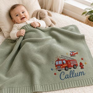 Può includere: Una coperta per neonati lavorata a maglia color verde salvia con il ricamo di un camion dei pompieri rosso, un elicottero, un idrante e piccole stelle. Il nome Callum è ricamato in corsivo blu scuro sotto il camion dei pompieri.