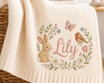 May include: A cream-colored knit baby blanket featuring an embroidered wreath design. The wreath includes a brown rabbit, a robin, a pink butterfly, and various flowers. The name Lily is embroidered in the center in a pink cursive script font.