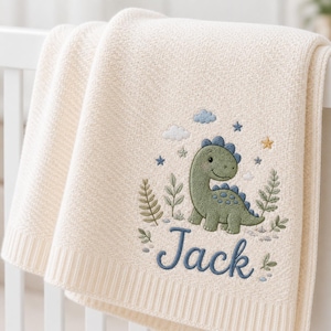 May include: A cream-colored knit blanket featuring an embroidered design of a green dinosaur surrounded by small plants, clouds, and stars. Below the dinosaur, the name Jack is embroidered in blue script lettering with a textured finish.