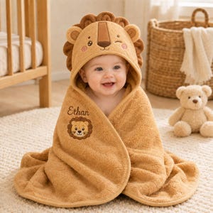 Op de afbeelding: Een beige babyhanddoek met capuchon, voorzien van een leeuwengezicht op de capuchon met oren en manen. De naam Ethan en een kleine leeuwengrafiek zijn op de voorzijde geborduurd. De stof heeft een zachte, getextureerde uitstraling over het geheel.