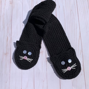 May include: Black knitted scarf with a cat face design. The scarf has two black button eyes, a pink button nose, and white yarn whiskers.