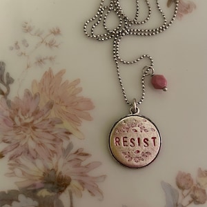 Op de afbeelding: Een zilveren ketting met een ronde hanger waarop "RESIST" in rode letters staat. De hanger is omgeven door een bloemmotief. De ketting heeft ook een kleine roze stenen hanger.
