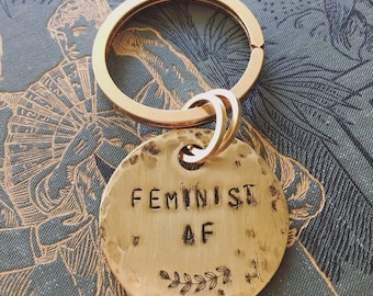 May include: Gold-toned keychain with a round, hammered metal tag that reads "FEMINIST AF" with a small leafy design below the text.