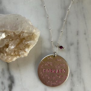 Puede incluir: Un collar de plata con un colgante redondo que dice "FORWARD" en letras rosas. El colgante está rodeado de un diseño floral. El collar tiene una pequeña cuenta de piedra preciosa púrpura.