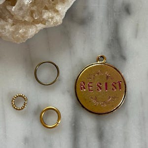 Op de afbeelding: Een verzameling sieradenonderdelen, waaronder een goudkleurige hanger met het woord "RESIST" in roze letters, een kleine goudkleurige ring, een getextureerde goudkleurige ring en een donkerbronzen ring. De items liggen op een marmeren oppervlak.