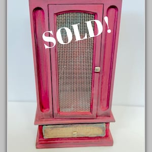 May include: A small, antique-style cabinet painted in a deep red hue, featuring a mesh-covered door and a small drawer. The word "SOLD!" is prominently displayed on the front. A pair of black tweezers with silver tips are at the bottom.