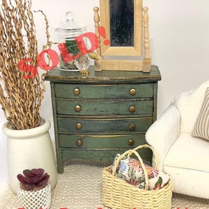 May include: Miniature green dresser with four drawers, gold knobs, and a small wooden mirror. A woven basket holds floral fabric, and a white vase contains dried branches. A small succulent is in a white pot. The text "Do No Buy - Sample work!" is visible.