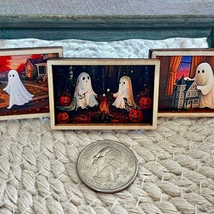 May include: Three miniature Halloween-themed paintings with wooden frames. Each painting depicts a ghost in a different scene: walking a dog, roasting marshmallows, and standing by a haunted house. A quarter is in the foreground.