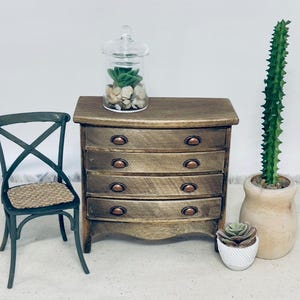 May include: Miniature wooden dresser with four drawers and copper-colored pulls. A small green chair with a woven seat is to the left. A glass jar with a lid contains rocks and a green plant. A tall cactus is in a wooden pot.