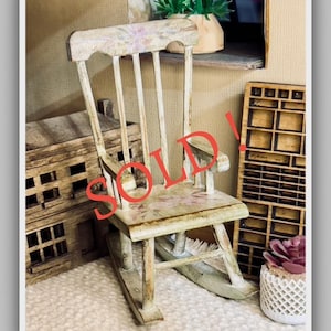May include: A miniature, distressed wooden rocking chair with floral accents. The chair is painted in shades of white and beige, with a weathered appearance. The word "SOLD" is written across the chair in red. The chair is part of a miniature scene.