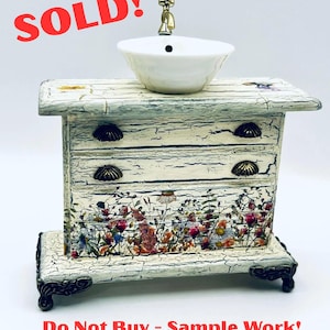 May include: Miniature decorative sink with a white ceramic bowl and a gold-colored faucet. The sink is set atop a distressed, off-white cabinet with floral accents and brass-toned hardware. The text "SOLD!" is visible.