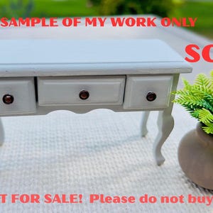 May include: A small, light gray desk with three drawers and curved legs. The desk has dark brown knobs and is next to a small brown vase with a green plant. The text "SAMPLE OF MY WORK ONLY" and "SOLD!" are visible.