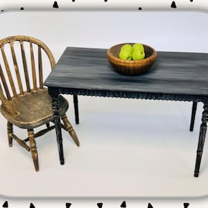 May include: A miniature wooden dining set with a distressed finish. The set includes a gray table with a decorative edge, a wooden bowl with two green apples, and a spindle-back chair. The table is approximately 10 inches long.