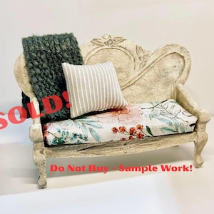 May include: Miniature antique-style sofa with floral and leaf patterned seat cushion, a striped pillow, and a dark green textured throw. The sofa is cream-colored with ornate carvings. Text on the image reads "SOLD!" and "Do Not Buy - Sample Work!"