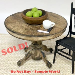 May include: Miniature round wooden table with a distressed finish, holding a wooden bowl of green apples, a white teacup, and a small book. A black chair is next to the table. The word "SOLD!" is written on the table.