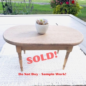 May include: A small, oval wooden table with four turned legs. A white ceramic pot with a succulent plant sits on the table. The word "SOLD!" is printed in red across the bottom of the table, with the text "Do Not Buy - Sample Work!" below it.