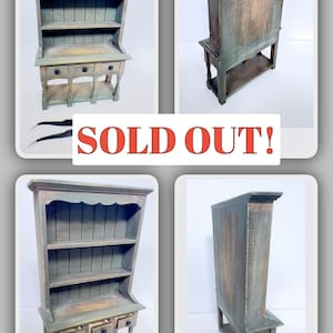 May include: A miniature wooden hutch painted in shades of gray and brown. The hutch features open shelves, three small drawers, and a scalloped top. The text "SOLD OUT!" is displayed in red.