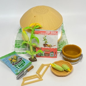 May include: A miniature gardening set featuring a corn stalk, a bag of Happy Gro Co seeds, a small shovel, and a woven basket with corn cobs. The set includes a yellow bucket, a hat, and a booklet titled "Corn Stalk".