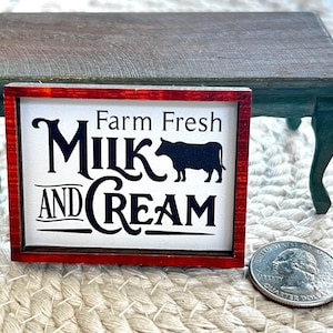May include: A miniature rectangular sign with a red frame and white background. The sign reads "Farm Fresh Milk and Cream" in black lettering, with a black cow silhouette. A U.S. quarter is next to the sign.