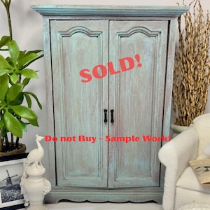 May include: A distressed, two-door cabinet painted in shades of teal and cream. The cabinet has black hardware and the word "SOLD!" is printed across the front in red. The text "Do not Buy - Sample Work!" is also visible.