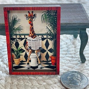 May include: Miniature framed artwork featuring a giraffe wearing glasses and reading a newspaper while sitting on a toilet. The artwork has a red frame and a black and white tiled floor background. The newspaper reads "NEE IONIN NO".
