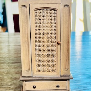 May include: A small, wooden cabinet with a light brown finish. The cabinet has a single door with a woven cane panel and a small drawer below. The cabinet is approximately 10 inches tall.