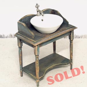 May include: Miniature bathroom sink with a white porcelain basin and a silver faucet. The sink is set on a distressed wooden vanity with a lower shelf. The vanity has a blue and gold finish. The text "SOLD!" and "Do Not Buy - Sample Work!" are visible.
