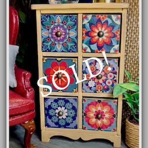 May include: A small, wooden cabinet with six drawers, each adorned with a colorful floral pattern. The drawers feature a variety of designs in shades of blue, orange, and pink. The word "SOLD!" is written across the front.